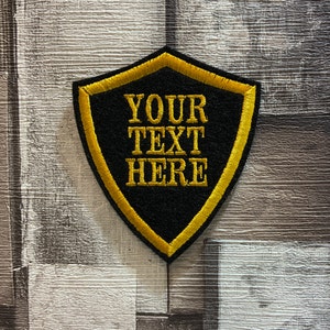 May include: Black embroidered patch shaped like a shield with a yellow border. The text "YOUR TEXT HERE" is embroidered in yellow on the patch.