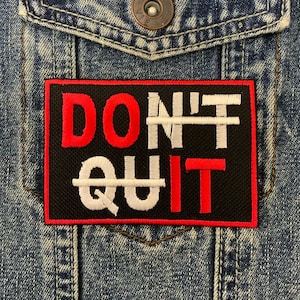May include: A black patch with white and red embroidered text that reads "DON'T QUIT" with lines through the "N" and the "Q". The patch is sewn onto a blue denim jacket.
