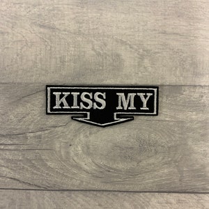 May include: Black and white embroidered patch with the text "KISS MY" in a white font on a black background. The patch has a white border and a white arrow pointing down.