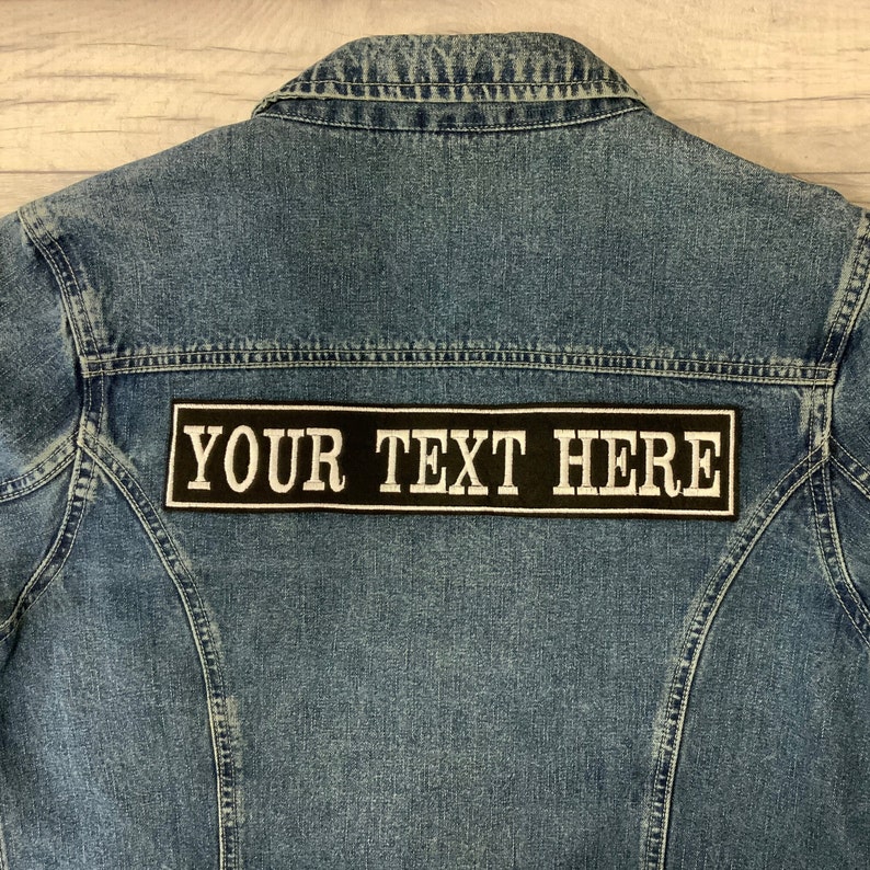 K&ouml;nnte beinhalten: Eine blaue Jeansjacke mit einem schwarzen und wei&szlig;en Patch, auf dem "YOUR TEXT HERE" steht.