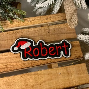 May include: A black embroidered patch with the name "Robert" in red, a white outline, and a Santa hat design. The patch is set against a wooden background, with festive decorations visible.
