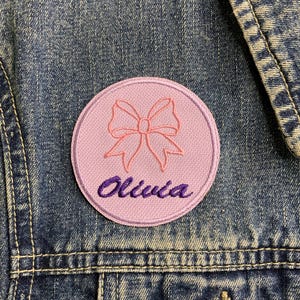 May include: A pink fabric patch with a pink bow and the name "Olivia" embroidered in purple thread.