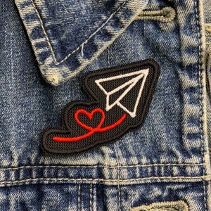 May include: A black and red embroidered patch featuring a white paper aeroplane and a red heart. The patch is sewn onto a blue denim jacket.
