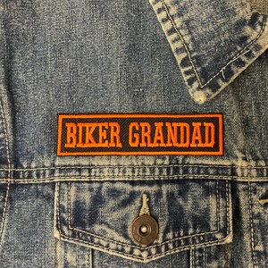 May include: A black and orange embroidered patch with the text "Biker Grandad" on a denim jacket.