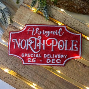 May include: Red and white embroidered patch with the text "Abigail North Pole Special Delivery 25 - Dec".