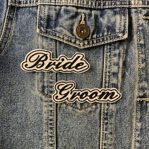 May include: Two white patches with black cursive lettering sewn onto a blue denim jacket. The patches say "Bride" and "Groom".
