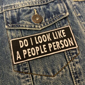 May include: A black patch with white text that reads "Do I look like a people person?" sewn onto a blue denim jacket.