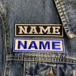 May include: Two embroidered patches with the word "NAME" in white lettering. The top patch is black with white stitching. The bottom patch is white with blue stitching.