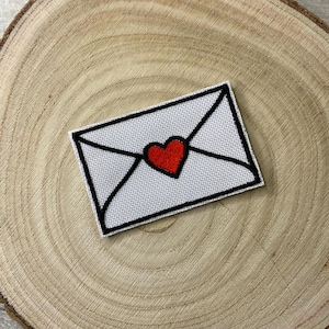 May include: White fabric envelope with a black outline and a red heart in the centre. The envelope is on a wooden surface.