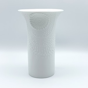 May include: White ceramic vase with a raised, embossed design of a flower. The vase has a flared top and a cylindrical body.