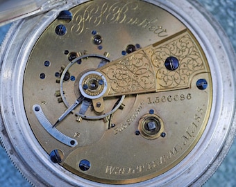 May include: Close-up of a vintage pocket watch mechanism. The brass-colored interior features intricate gears, screws, and a decorative gold-toned plate with engraved details. The watch is marked "WALTHAM, MASS." and includes the serial number 1,566,696.