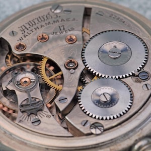 May include: Detailed view of a vintage pocket watch mechanism. The watch face is marked with "A.W. Co. Waltham, Mass." and "17 Jewels." The intricate gears and components are visible, highlighting the watch's internal workings. The watch has a silver-coloured metal casing.