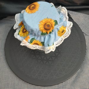 May include: A circular jar lid cover made of light blue fabric featuring a yellow sunflower pattern with green leaves. The cover is gathered with an elastic band and finished with a delicate white lace trim around the entire outer edge.