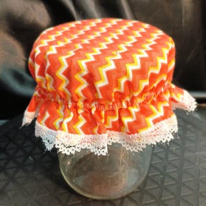May include: A clear glass jar topped with a fabric cover featuring a vibrant orange, yellow, and white chevron pattern. The fabric is gathered with an elastic band and finished with a delicate white lace trim along the bottom edge.