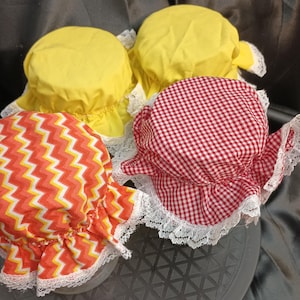 May include: Four jar covers made of fabric, featuring two solid yellow covers, one red and white gingham checkered cover, and one orange, yellow, and white chevron patterned cover. Each cover is finished with a white lace trim edge.
