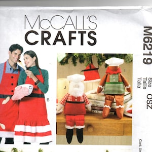 May include: McCall's Crafts pattern featuring Christmas-themed aprons and decorative figures. The aprons are red with black and white accents, and the figures include a Santa Claus and a gingerbread man, all with chef hats.