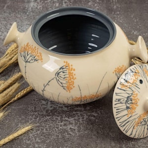 Puede incluir: Cazuela de cerámica color crema con tapa, con un diseño floral en naranja y gris. El plato tiene un interior gris oscuro y asas. Espigas de trigo dispuestas alrededor del plato sobre una superficie gris.
