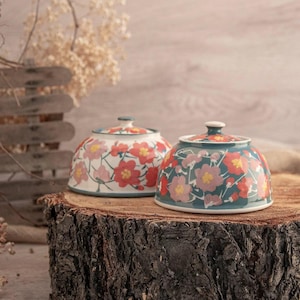 Op de afbeelding: Twee keramische kommen met deksels en bloempatronen. Eén kom heeft een witte achtergrond met rode en roze bloemen, de andere heeft een teal achtergrond met roze en rode bloemen. Beide kommen staan op een boomstronk.