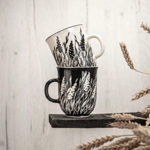 Puede incluir: Dos tazas de cerámica apiladas con un diseño floral en blanco y negro. La taza inferior es negra con flores blancas, y la taza superior es blanca con flores negras. Ambas tazas tienen un asa curva y están sobre una estantería de madera oscura.