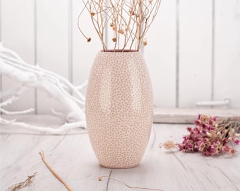 Może przedstawiać: Jasnoróżowy ceramiczny wazon w kropki, wypełniony suchymi gałązkami z małymi strąkami nasiennymi. Owalny wazon stoi na jasnej drewnianej powierzchni. W tle widoczne są suszone kwiaty.