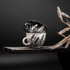 May include: Two stacked ceramic teacups with a black and white floral design. The bottom cup is white with black floral accents, and the top cup is black with white floral accents. The cups have curved handles and sit on a dark wooden shelf.