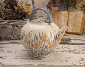 May include: Cream ceramic teapot with a blue handle and spout. The teapot is decorated with a pattern of grey blades of grass and orange floral accents. The lid also has a matching design.