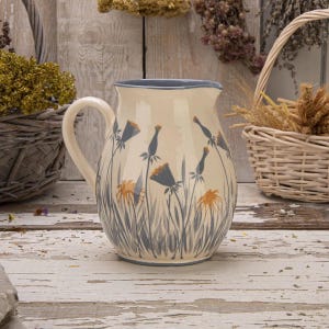 Op de afbeelding: Crème keramische kan met een blauwe rand en handvat. De kan is versierd met een bloemenontwerp met grijze en oranje bloemen en hoog grijs gras. De kan staat op een wit houten oppervlak.