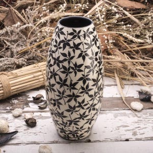 Op de afbeelding: Een hoge, crèmekleurige keramische vaas met een zwart bladerpatroon. De vaas heeft een brede body die naar boven toe smaller wordt en een zwart interieur. De decoratieve vaas is geschikt voor het presenteren van bloemen of als op zichzelf staand accentstuk.
