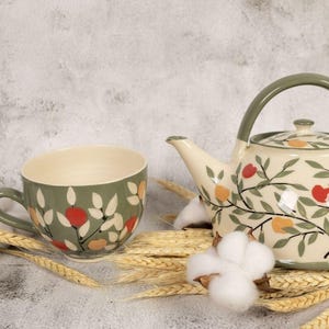 Puede incluir: Un juego de té de cerámica con una tetera y una taza. La tetera y la taza están decoradas con un estampado floral de flores rojas, naranjas y blancas sobre un fondo verde. La tetera tiene un asa y una tapa verdes.