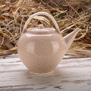 May include: A ceramic teapot with a light pink base color and a pattern of small white dots. The teapot has a curved spout, a rounded body, and a white handle. The lid has a small knob. The teapot is on a white wooden surface.