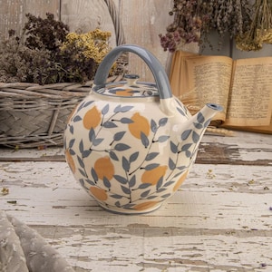 Peut inclure: Une théière en céramique avec un corps rond, une anse grise et un bec verseur. La théière est décorée de citrons jaunes et de feuilles grises sur un fond crème. Le couvercle a un petit bouton. La théière est posée sur une surface en bois blanc.
