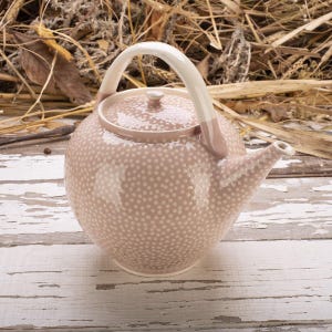 May include: A pink ceramic teapot with a white handle and lid. The teapot is covered in small white polka dots. The handle and the base of the handle are a light pink color. The teapot has a spout for pouring.