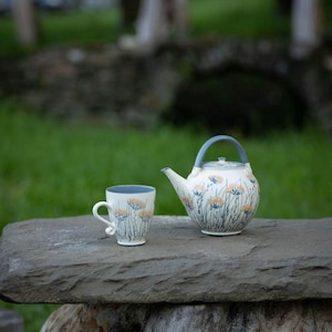 Peut inclure: Un service à thé en céramique comprenant une tasse et une théière avec un motif floral. La tasse et la théière sont blanches avec un bord et une anse bleus, et décorées de motifs floraux orange et bleus. L'ensemble est posé sur une surface en pierre avec un arrière-plan vert flou.
