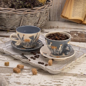 May include: Two ceramic teacups and saucers with a floral design in orange and white on a grey-blue background. One cup is filled with coffee beans. The cups sit on a decorative tray with sugar cubes and a spoon.