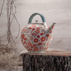 Può includere: Teiera in ceramica con motivo floreale. La teiera presenta un corpo arrotondato, un manico turchese e un beccuccio. Il design include fiori rossi e rosa su base bianca. Poggia su una superficie di legno.