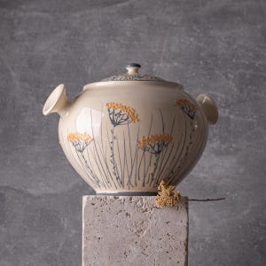 Puede incluir: Una fuente de cerámica crema con tapa, con un diseño floral en naranja y azul. La fuente tiene dos asas y se apoya sobre un pedestal de piedra. La tapa tiene un pequeño pomo azul y un borde con dibujos.