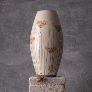 Könnte beinhalten: Cremefarbene Keramikvase mit dunkelblauem Rand und Boden. Die Vase hat ein Design aus hohen, dünnen schwarzen Linien und gelb-orangen Blütenclustern. Sie wird auf einem Stapel hellgrauer Steinblöcke ausgestellt. Die Vase ist etwa 25 cm hoch.