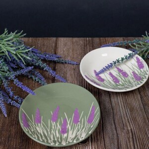 Könnte beinhalten: Zwei kleine Keramikschalen mit Lavendeldesign. Eine Schale ist hellgrün, die andere weiß. Beide haben lila Lavendelzweige und grünes Gras aufgemalt. Lavendelzweige sind auf einer Holzoberfläche um die Schalen angeordnet.