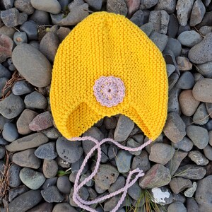 Può includere: Un cappello di lana giallo con un fiore rosa in cima e una stringa di chiusura rosa. Il cappello è appoggiato su un letto di pietre grigie.