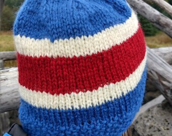 May include: A blue, red, and white knit beanie hat. The hat features a blue crown, a white stripe, and a red stripe. The hat is made of a thick, textured yarn and appears to be handmade. The hat is worn by a person.