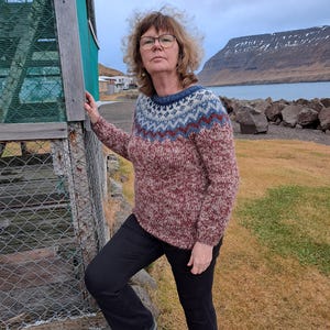 May include: A woman wearing a red, white, and blue patterned sweater with a brown background. She is standing next to a wooden structure with a wire mesh fence. The woman is looking at the camera.