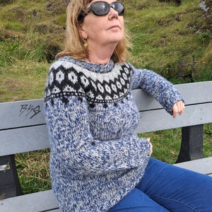 May include: A woman wearing a blue and white patterned sweater with a black and white geometric design. She is sitting on a wooden bench and looking up.