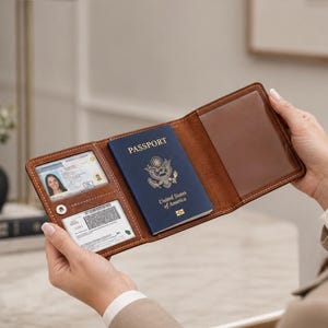以下が含まれることがあります： ステッチが施された三つ折りデザインの茶色のレザー製パスポートケース。内側には身分証明書用の透明なプラスチック窓、カード用スロット、そして中央には青いアメリカ合衆国のパスポートを収納するスリーブが備わっています。