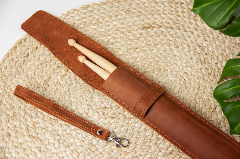 May include: A brown leather drumstick holder with a strap and a metal clasp. Two wooden drumsticks are inside the holder.