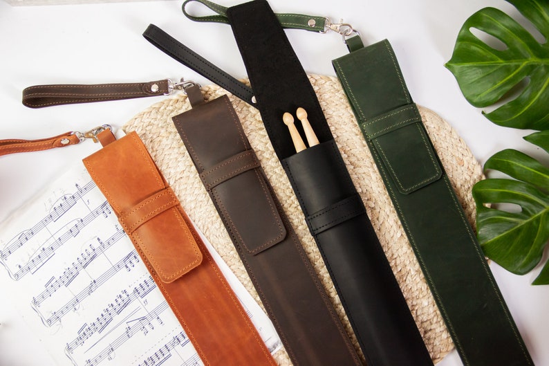 May include: Five leather drumstick holders in different colours, including brown, black, green, and orange. Each holder has a strap and a pocket for the sticks.