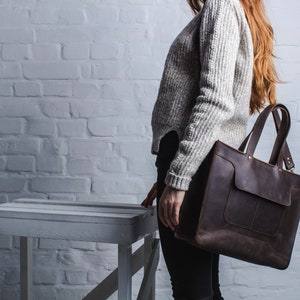 K&ouml;nnte beinhalten: Eine braune Ledertasche mit einer gro&szlig;en Tasche auf der Vorderseite. Die Tasche hat zwei obere Griffe und einen Schultergurt.