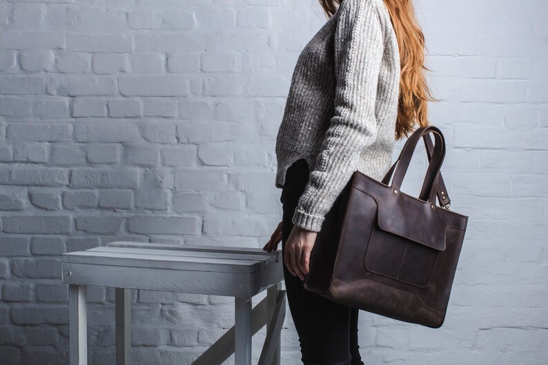 K&ouml;nnte beinhalten: Eine braune Ledertasche mit einer gro&szlig;en Tasche auf der Vorderseite. Die Tasche hat zwei obere Griffe und einen Schultergurt.