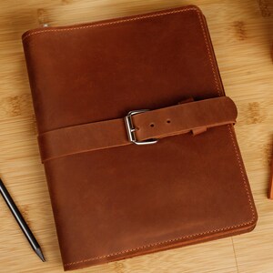 May include: A brown leather journal with a buckle closure. The journal is on a wooden surface.