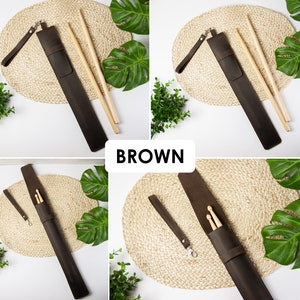 May include: Brown leather drumstick holder with a strap and two wooden drumsticks. The holder is on a woven mat with green leaves in the background.
