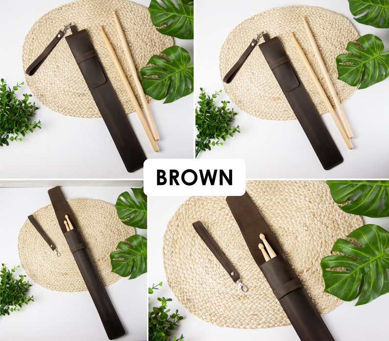 May include: Brown leather drumstick holder with a strap and two wooden drumsticks. The holder is on a woven mat with green leaves in the background.